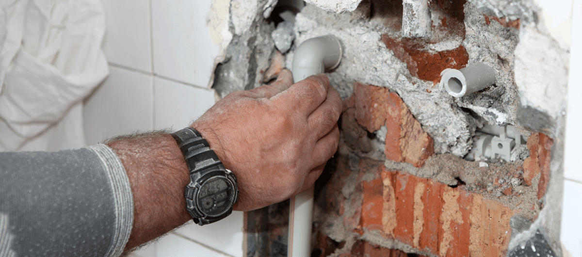 Egy férfi kéz fogja a bontott falból kiálló vízvezetéket csőtörés elhárítása közben.